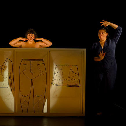 photo du spectacle Filles et soie pour toutes et tous où on voit une femme nue derrière un grand carton avec des vêtements dessinés à côté d'une femme qui traduit en langue des signes