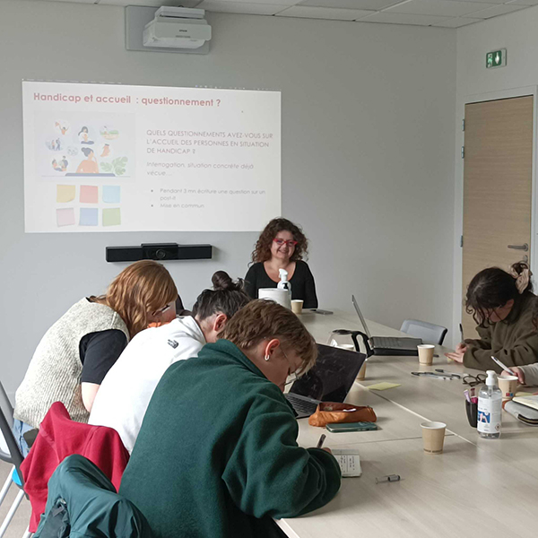 plusieurs personnes dans une salle assises autour d'une table, devant une femme en fauteuil électrique qui sourit