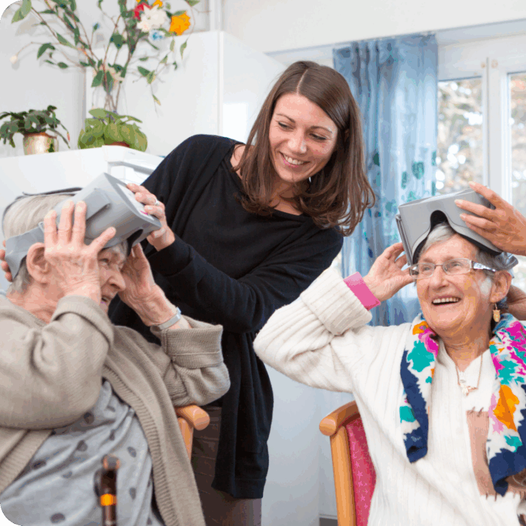 2 personnes agées qui enlèvent leur casque de réalité virtuelle, une femme brune et derrière eux, elle les aide, ils sourient