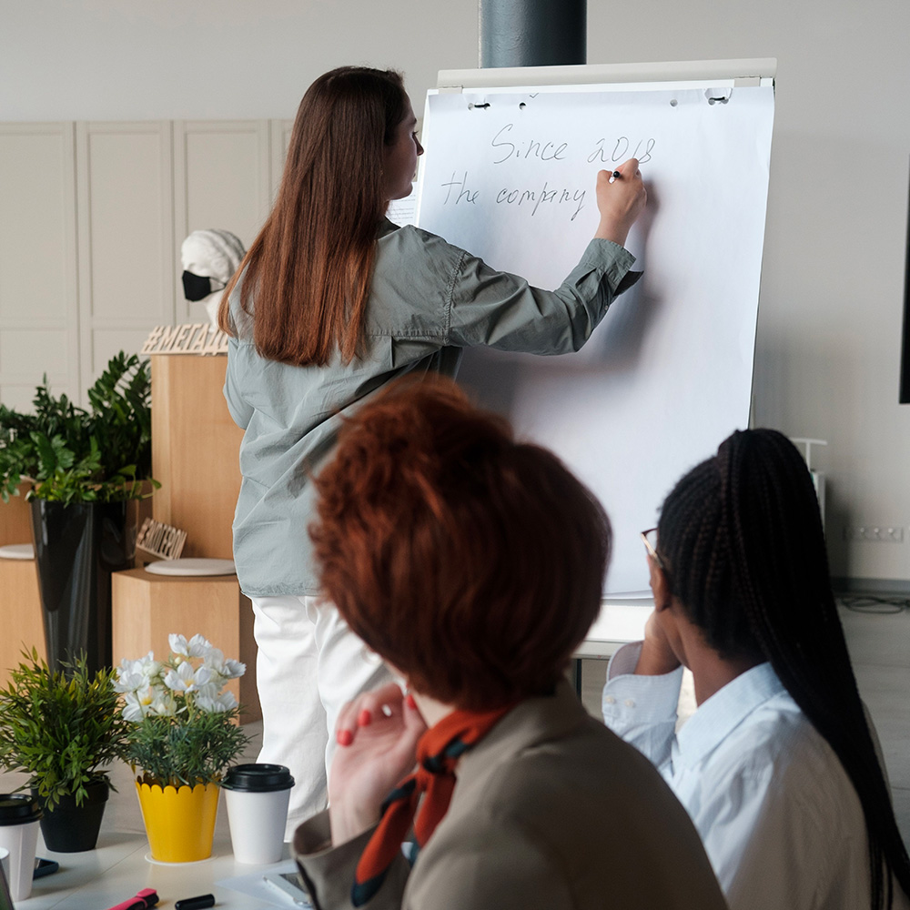 photo de 3 personnes, une écrit sur un paperboard, 2 autres sont assises autour d'un bureau